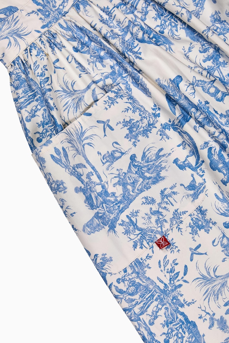 May include: White and blue toile fabric with a pocket. The fabric features a detailed pastoral scene with figures, trees, and birds. A red tag with a floral design is visible on the pocket. The overall design is reminiscent of classic French country style.