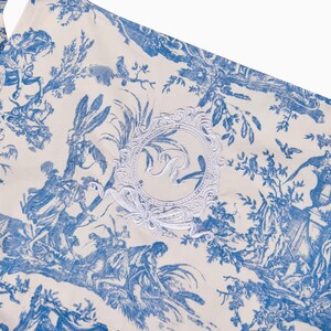 May include: Close-up of a cream-colored fabric with a blue toile pattern depicting classical scenes. A white embroidered emblem with the letter "R" is centered on the fabric. The fabric has a ruffled edge.
