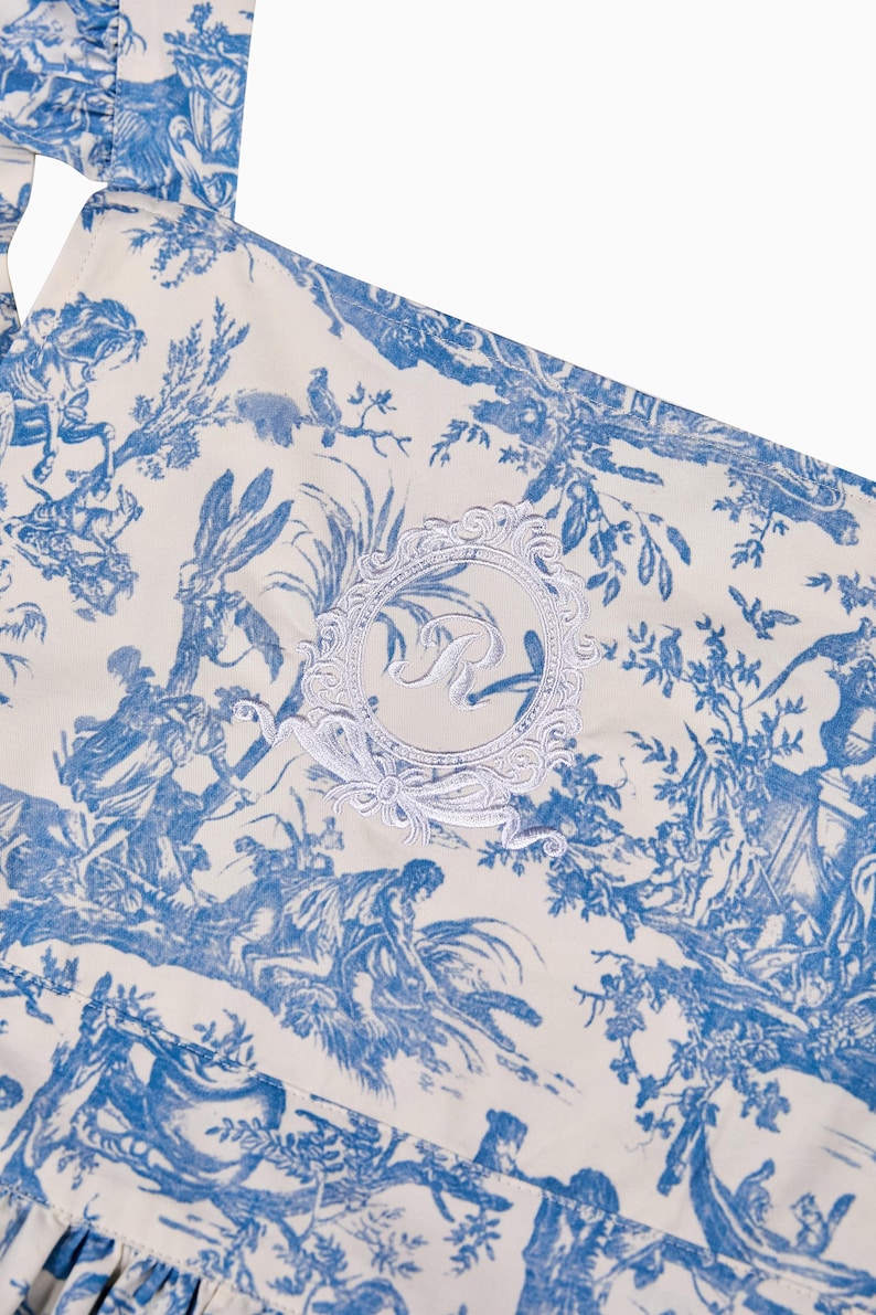 May include: Close-up of a cream-colored fabric with a blue toile pattern depicting classical scenes. A white embroidered emblem with the letter "R" is centered on the fabric. The fabric has a ruffled edge.