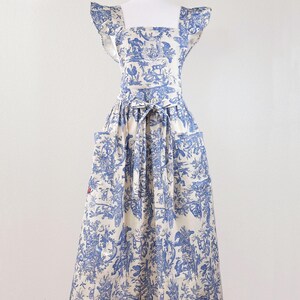 May include: A blue and white apron with a toile pattern, featuring a square neckline, ruffled sleeves, and two front pockets. The apron is tied at the waist with a matching fabric belt. The fabric has a cream background with blue illustrations.