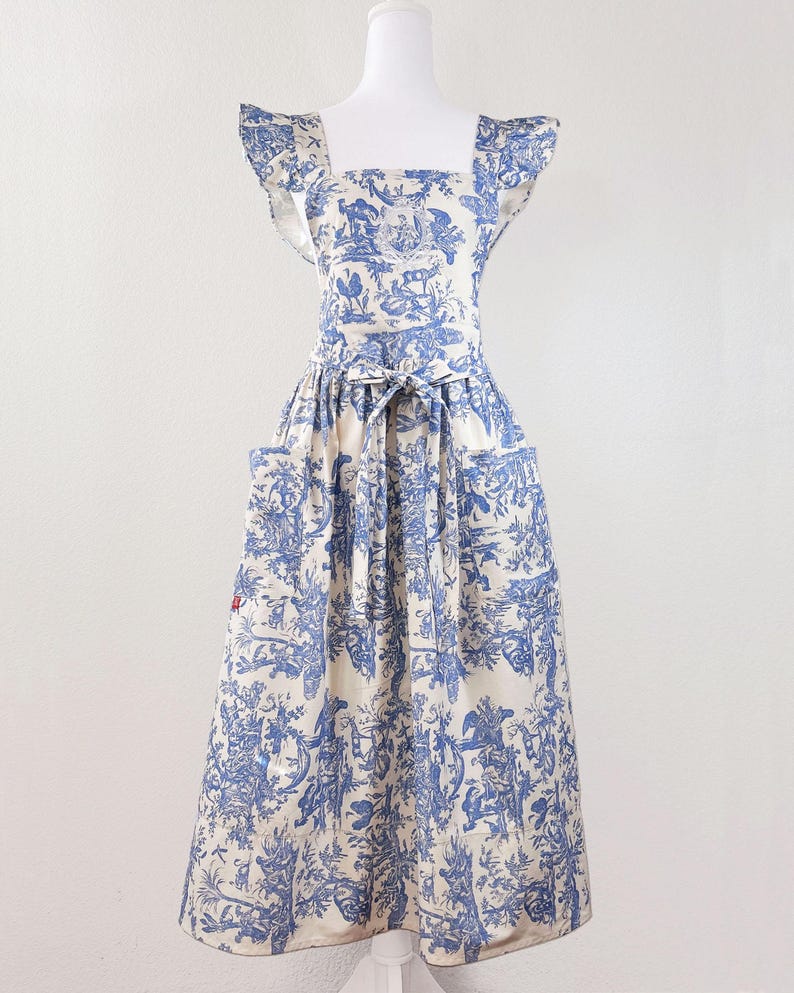May include: A blue and white apron with a toile pattern, featuring a square neckline, ruffled sleeves, and two front pockets. The apron is tied at the waist with a matching fabric belt. The fabric has a cream background with blue illustrations.