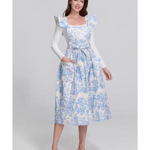 May include: A blue and white apron dress with a toile pattern. The dress has a square neckline, ruffled shoulder details, and a tie waist. It is worn over a white long-sleeved shirt. The dress has pockets and is knee-length.