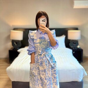 May include: A blue and white floral print apron with a matching tie and pockets. The apron is worn over a light blue long-sleeved shirt. The apron has a ruffled trim on the shoulders.