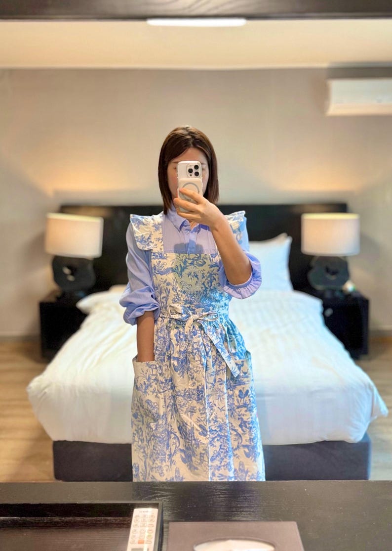May include: A blue and white floral print apron with a matching tie and pockets. The apron is worn over a light blue long-sleeved shirt. The apron has a ruffled trim on the shoulders.