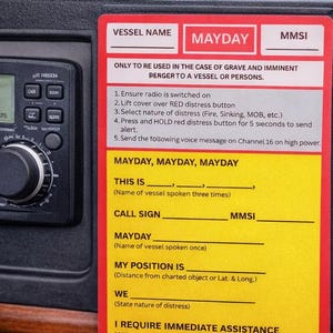 May include: Marine radio with a digital display showing channel 16, alongside a "Mayday" distress procedure card. The card provides instructions for emergency calls, with fields for vessel name, call sign, and position.