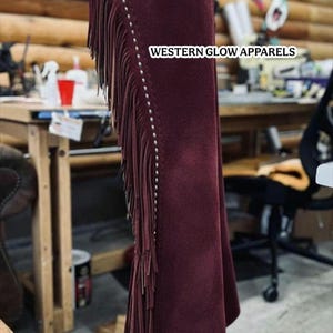 Op de afbeelding: Bordeaux suède chaps met decoratieve leren accenten en franjes. De chaps hebben een bloemmotief op de leren afwerking en zijn versierd met zilverkleurige studs. De tekst "WESTERN GLOW APPARELS" is zichtbaar.