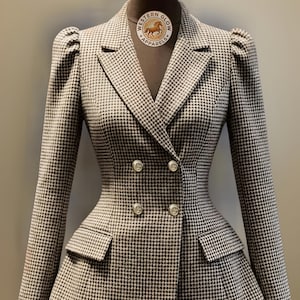 May include: A houndstooth blazer with a double-breasted design. The jacket has gold buttons, a fitted waist, and small pockets. The label on the mannequin reads "WESTERN GLOW APPARELS".
