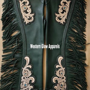 May include: Dark green leather chaps featuring long side fringe, silver-tone metal studs, and light tan floral tooling with small turquoise accents. The text Western Glow Apparels is printed in white in the centre of the garment.
