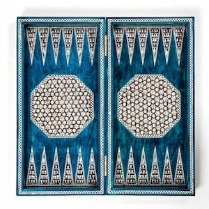 Puede incluir: Un tablero de backgammon azul con un diseño incrustado decorativo. El tablero presenta un patrón octogonal central y puntas triangulares, todo enmarcado con un borde blanco y negro. El tablero está articulado y parece cerrado.