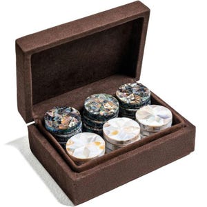 Könnte beinhalten: Eine braune Samtbox mit sechs dekorativen runden Untersetzern. Die Untersetzer haben ein Mosaikdesign mit schillernden und weißen Muschelteilen. Die Box ist geöffnet und zeigt die Untersetzer.