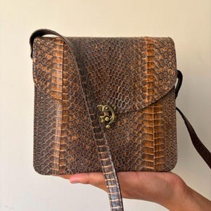 May include: A square-shaped crossbody bag featuring a dark brown and tan snakeskin-like textured pattern. The bag has a fold-over flap secured with a brass-toned metal clasp and includes a matching long shoulder strap for carrying.