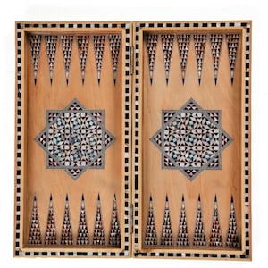 May include: An open, hinged backgammon board with detailed inlaid patterns. The light wood surface is adorned with geometric designs in black, white, and red. A central star motif and triangular points contribute to the board's decorative appearance.