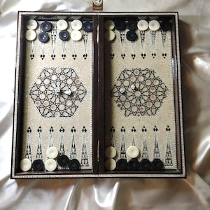 May include: A wooden backgammon board with a decorative inlaid design. The board is open, revealing a playing surface with alternating black and white points. Black and white game pieces are arranged on the board, with dice visible in the center.