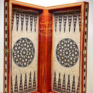 Puede incluir: Un tablero de backgammon de madera con intrincados diseños incrustados. El tablero presenta un marco naranja cálido con patrones geométricos blancos y negros. El centro tiene un diseño circular y texto árabe.