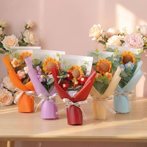 May include: Five colorful crochet flower bouquets wrapped in paper and tied with white ribbon. Each bouquet features a variety of crocheted flowers, including sunflowers and other blooms, in shades of orange, purple, red, yellow, and blue.