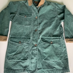 May include: A green jacket with a brown collar and cuffs. The jacket has four front pockets, a button-up closure, and brown buttons. The jacket is made of a durable fabric and is suitable for outdoor wear.