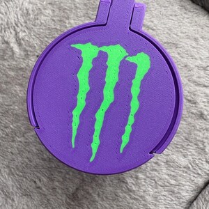 K&ouml;nnte beinhalten: Ein runder, violett 3D-gedruckter Beh&auml;lter mit einem leuchtend gr&uuml;nen Monster Energy Logo auf dem Deckel. Der Deckel hat eine kleine Lasche zum &Ouml;ffnen, und das gesamte Objekt weist die f&uuml;r die additive Fertigung typische geschichtete Textur auf.