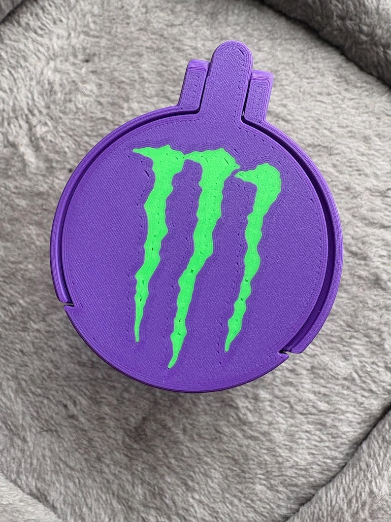 K&ouml;nnte beinhalten: Ein runder, violett 3D-gedruckter Beh&auml;lter mit einem leuchtend gr&uuml;nen Monster Energy Logo auf dem Deckel. Der Deckel hat eine kleine Lasche zum &Ouml;ffnen, und das gesamte Objekt weist die f&uuml;r die additive Fertigung typische geschichtete Textur auf.