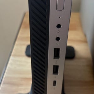 May include: A black and silver desktop computer in a vertical orientation. The front panel has a power button, audio jacks, and USB ports. The side panel has a textured design. The computer is in a black stand.