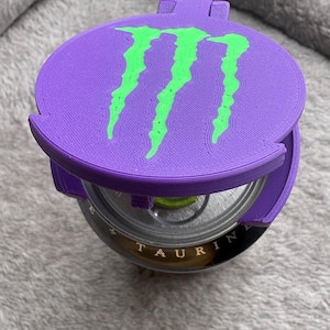 K&ouml;nnte beinhalten: Ein violetter, 3D-gedruckter Kunststoffdeckel f&uuml;r Getr&auml;nkedosen mit einem leuchtend gr&uuml;nen Monster Energy-Krallenlogo auf der Oberseite. Der runde Deckel liegt auf einer Metalldose, wobei das Wort TAURINE am Dosenrand sichtbar ist.