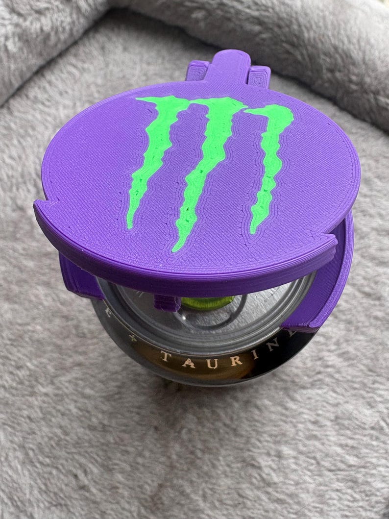K&ouml;nnte beinhalten: Ein violetter, 3D-gedruckter Kunststoffdeckel f&uuml;r Getr&auml;nkedosen mit einem leuchtend gr&uuml;nen Monster Energy-Krallenlogo auf der Oberseite. Der runde Deckel liegt auf einer Metalldose, wobei das Wort TAURINE am Dosenrand sichtbar ist.
