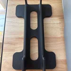 以下が含まれることがあります： 長方形のベースと2つの垂直サポートを備えた、黒色の3Dプリントスタンド。スタンドには2つの楕円形の切り欠きと、上部に2つの隆起したセクションがあります。スタンドは、アイテムを直立させて保持するように設計されています。