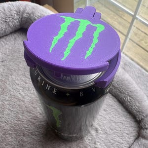 K&ouml;nnte beinhalten: Ein violetter, 3D-gedruckter Kunststoff-Dosenaufsatz mit einem leuchtend gr&uuml;nen Monster Energy-Logo auf der Oberseite. Das kreisf&ouml;rmige Zubeh&ouml;rteil sitzt fest auf dem silbernen Verschluss einer schwarzen Aluminium-Getr&auml;nkedose mit Text.