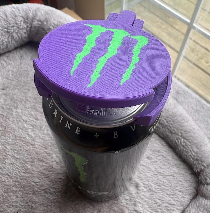 K&ouml;nnte beinhalten: Ein violetter, 3D-gedruckter Kunststoff-Dosenaufsatz mit einem leuchtend gr&uuml;nen Monster Energy-Logo auf der Oberseite. Das kreisf&ouml;rmige Zubeh&ouml;rteil sitzt fest auf dem silbernen Verschluss einer schwarzen Aluminium-Getr&auml;nkedose mit Text.