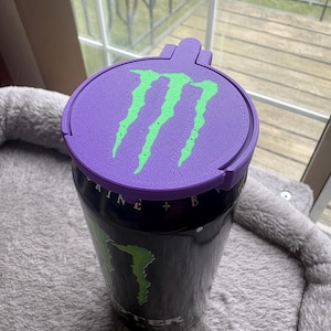 K&ouml;nnte beinhalten: Eine schwarze Aluminium-Getr&auml;nkedose mit einem individuellen, 3D-gedruckten violetten Kunststoffdeckel. Der Deckel zeigt ein leuchtend gr&uuml;nes, erhabenes Monster Energy-Logo. Die Dose selbst tr&auml;gt das teilweise sichtbare Monster Energy-Branding.