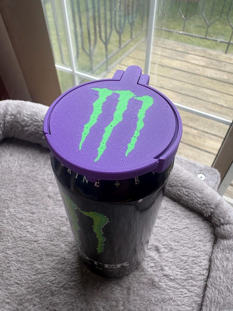 K&ouml;nnte beinhalten: Eine schwarze Aluminium-Getr&auml;nkedose mit einem individuellen, 3D-gedruckten violetten Kunststoffdeckel. Der Deckel zeigt ein leuchtend gr&uuml;nes, erhabenes Monster Energy-Logo. Die Dose selbst tr&auml;gt das teilweise sichtbare Monster Energy-Branding.