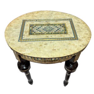 May include: A round side table featuring an intricate inlay of cream-colored mother-of-pearl tiles across the top and apron. The center of the tabletop displays a rectangular geometric pattern with blue and gold accents. Two dark brown legs support it.