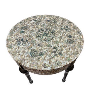 May include: A round side table featuring a tabletop and apron covered in a mosaic of iridescent mother-of-pearl tiles. The surface displays a central floral motif surrounded by scrolling leaf patterns in shades of white, cream, and grey.