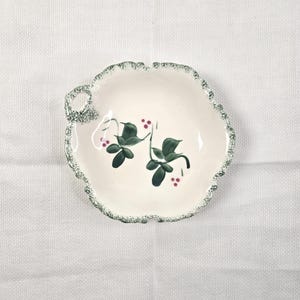 May include: A small, off-white ceramic dish with a scalloped edge and a small handle. The dish features a painted design of green leaves and red berries. The rim is decorated with a green speckled pattern.