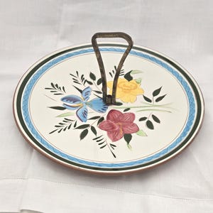 May include: A round serving platter with a floral design. The platter has a cream-coloured surface with a blue, green, and brown border. The floral design includes blue irises, yellow roses, and red flowers. A metal handle is attached.