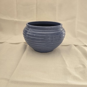 May include: A round, blue ceramic planter with a ribbed exterior. The planter has a wide opening and a slightly curved shape. The color is a muted blue, and the texture appears smooth. The planter is suitable for indoor or outdoor use.