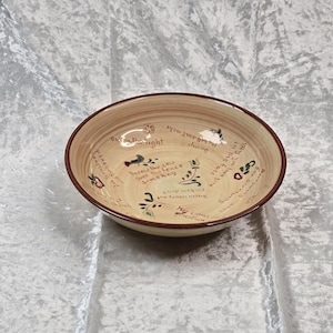 May include: A beige ceramic bowl with a brown rim and a cream-colored interior. The bowl features handwritten text and small illustrations in various colors, including red, green, and blue. The text includes phrases like "Throw the cow over the fence somehow."