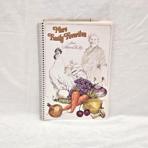 May include: A spiral-bound cookbook titled More Family Favorites from Miriam B. Loo. The cover features a line-art illustration of a person, a rooster, and a colorful arrangement of produce including grapes, carrots, an onion, and cherries.
