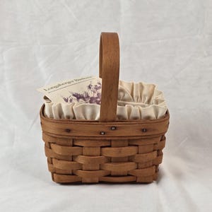 Puede incluir: Una cesta rectangular de madera trenzada marrón con un asa vertical única y un forro de tela color crema con borde con volantes. Una pequeña tarjeta rectangular con el texto Longaberger Baskets y una ilustración floral morada está insertada.