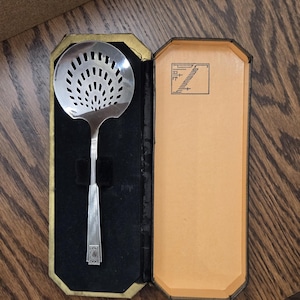 May include: A silver-colored slotted spoon with a decorative handle, presented in a black velvet-lined case with a gold trim. The case is open, revealing the spoon and a tan interior. The spoon has a circular head with multiple holes.