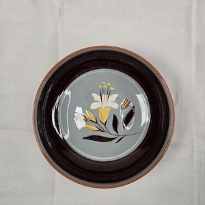 May include: Round ceramic plate with a floral design. The plate features a light blue center with a white and yellow flower and brown and black accents. The rim is dark brown with a light brown edge.