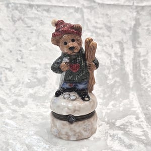 May include: A decorative trinket box featuring a brown bear figurine wearing a red hat and green sweater with a heart design. The bear holds skis and stands atop a snow-covered base. The box is beige with a floral pattern.