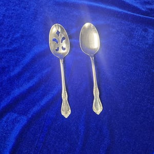 Puede incluir: Dos cucharas de metal plateado con mangos ornamentados. Una cuchara presenta un patrón recortado decorativo en el cuenco que recuerda a una flor de lis, mientras que la otra es una cuchara de servir sólida con detalles a juego.