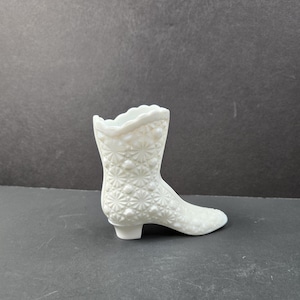 Op de afbeelding: Een witte, decoratieve vaas in de vorm van een laars. De vaas is gemaakt van ondoorzichtig glas met een textuur van een sterrenpatroon. De laars heeft een kleine hak en een geschulpte bovenrand. Het object staat tegen een donkere achtergrond.