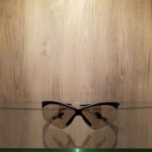 May include: Black-framed safety glasses with tinted lenses. The glasses are resting on a clear glass shelf, with a wooden background. The lenses reflect a hand.