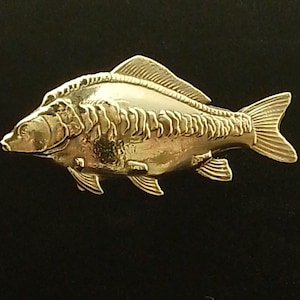 Op de afbeelding: Een goudkleurige vissenpin met gedetailleerde schubben, vinnen en een staart. De pin is geplaatst tegen een zwarte achtergrond, wat de metalen glans benadrukt. Deze decoratieve pin is een uniek accessoire.