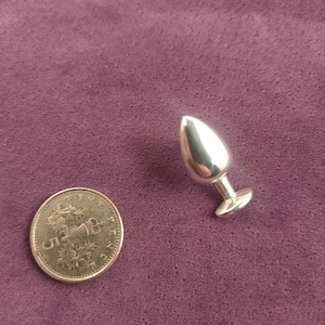 Puede incluir: Un tapón de metal plateado con una cabeza en forma de lágrima y una base redonda. El tapón es aproximadamente del tamaño de una moneda británica de cinco peniques.