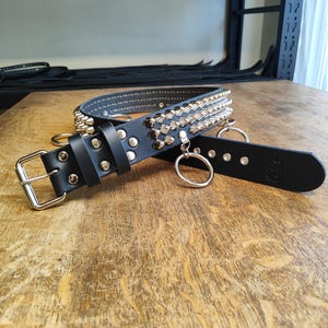 Puede incluir: Collar de cuero negro con tachuelas cónicas y remaches de color plateado. El collar presenta una hebilla plateada y dos anillos plateados. El collar está sobre una superficie de madera.