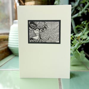 Op de afbeelding: Een crèmekleurige wenskaart met een zwart-wit illustratie. Het kunstwerk toont een gestileerde figuur met ingewikkelde patronen en bloemendetails in een zwart rechthoekig kader. De kaart wordt getoond op een lichtgroen oppervlak.