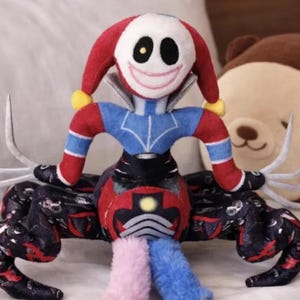May include: A soft plush toy of a jester-like figure with a red and blue outfit, white face, and a smiling expression. The toy has a black and red body with spider-like legs and colourful accents. The plush is detailed.