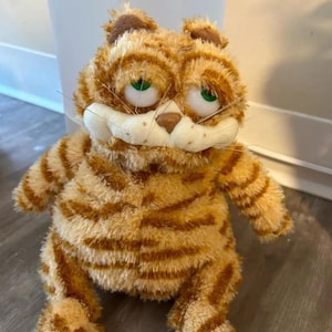 May include: A plush orange cat toy with brown stripes, green eyes, and a white muzzle. The soft toy has a slightly grumpy expression and is in a sitting position. The cat has a soft, furry texture and is a popular cartoon character.